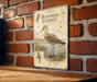 A vintage-style metal sign featuring illustrations of a bird, likely a stork, standing in a grassy area with a brick wall in the background.