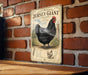 A vintage-style metal sign featuring an illustration of a black chicken, mounted on a brick wall.