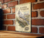 A metal sign with illustrations and text is mounted on a brick wall, depicting a juniper titmouse perched on a branch.