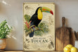A vintage-style metal sign featuring a colorful toucan perched on a branch, surrounded by tropical foliage and plants, with a wooden cutting board and lemons in the foreground.
