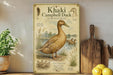 a poster of a khaki Campbell duck, with illustrations and text providing information about the bird. The poster is displayed on a shelf next to a wooden cutting board and a potted plant.