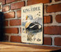 A metal sign with a detailed illustration of a king eider, a type of sea bird, is mounted on a brick wall.