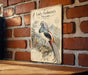 A vintage-style metal plaque featuring an illustration of a colorful bird, likely a peacock, perched on a branch against a brick wall background.