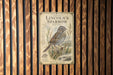 a vintage-style metal plaque with an illustration of a Lincoln's Sparrow, a small bird with a brown and white coloration, perched on a branch. The plaque is mounted on a wooden wall with vertical slats, creating a rustic and natural backdrop for the artwork.