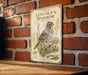 A vintage-style metal sign featuring an illustration of a Lincoln's Sparrow, mounted on a brick wall.