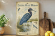 a vintage-style illustration of a blue heron, with a wooden paddle and a potted plant in the background.