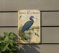 A metal sign featuring an illustration of a blue heron, with text and bird illustrations, mounted on a wall next to a plant.