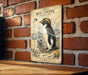 a vintage-style metal plaque with an illustration of a macaroni penguin, mounted on a brick wall.