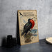a vintage-style sign with illustrations of a frigatebird, a large seabird known for its distinctive red head and black body. The sign is placed on a table next to a stack of black camera lenses and a white bowl.