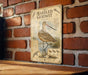 a vintage-style metal plaque with an illustration of a bird, specifically a stork, standing on a brick wall.
