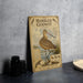 a vintage-style metal plate with an illustration of a bird, specifically a sandpiper, standing on a beach. The plate is placed on a countertop next to some black camera lenses.