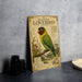 A framed illustration of a masked lovebird, a colorful parrot species, is displayed on a table next to a stack of black camera lenses.