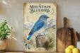 a vintage-style poster of a bluebird perched on a tree stump, surrounded by pine cones and pine needles, with a mountain backdrop.