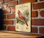 A vintage-style metal sign featuring a red cardinal perched on a pine branch, with pine cones and a map of the United States in the background.