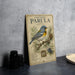 A vintage-style metal plaque with a bird illustration and text, placed on a gray surface next to some black camera lenses.