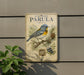 a vintage-style sign for the bird species Parula, which is a Northern Parula. The sign displays a detailed illustration of the bird perched on a branch with pine cones and berries. The sign is mounted on a wall, and there is a plant visible to the left of the sign.