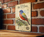 a vintage-style metal sign with a colorful illustration of a male bird, likely a painted bunting, perched on a branch. The sign is mounted on a brick wall and appears to be a decorative piece.
