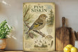 a vintage-style illustration of a pine siskin bird, with a detailed depiction of its features and habitat. The illustration is displayed on a wooden surface, accompanied by a wooden paddle and a potted plant.
