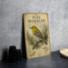A vintage-style metal sign with illustrations of pine warblers and pine needles, placed on a gray countertop next to a stack of black camera lenses.