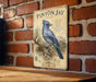 a vintage-style illustration of a blue bird, likely a piny jay, perched on a branch against a brick wall background.