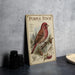 a vintage-style metal sign with a detailed illustration of a purple finch perched on a branch, surrounded by pine cones and berries.