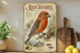 a vintage-style illustration of a red crossbill bird perched on a pine branch, surrounded by pine cones and pine needles. The bird is the main focus of the image, which is set against a backdrop of pine trees and mountains. The illustration is framed by a decorative border, and the text "The red crossbill" is prominently displayed at the top.