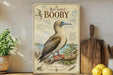 a vintage-style illustration of a red-footed booby, a type of seabird, perched on a branch with a birdhouse design. The illustration is accompanied by text that provides information about the red-footed booby, including its habitat, diet, and conservation status. The image is displayed on a shelf, with a potted plant, a wooden cutting board, and some lemons nearby.