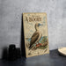 a vintage-style metal sign with a detailed illustration of a red-footed booby, a type of seabird, perched on a branch. The sign is placed on a gray countertop, with a stack of black camera lenses and a white dish visible in the background.