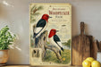 a vintage-style illustration of a pair of red-headed woodpeckers perched on a tree branch, surrounded by pine needles and flowers. The illustration is set against a backdrop of a natural landscape, with a potted plant and a wooden cutting board visible in the foreground.