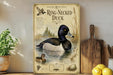 A framed illustration of a ring-necked duck is displayed on a shelf, accompanied by a wooden cutting board and a potted plant.