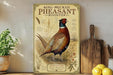 A framed illustration of a ring-necked pheasant rooster is displayed on a shelf, surrounded by kitchen utensils and a potted plant.