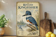 a framed illustration of a blue kingfisher bird, with a detailed illustration of the bird's features and habitat. The frame is placed on a wooden surface, with a potted plant and a wooden cutting board visible in the background.