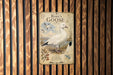 A vintage-style metal plaque featuring a white goose, with a map and text below it, mounted on a wooden wall.