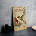 A vintage-style metal sign depicting a hummingbird, with text and illustrations, is displayed on a gray countertop next to a stack of black camera lenses.