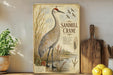 A framed illustration of a sandhill crane is displayed on a shelf, surrounded by various kitchen utensils and a potted plant.