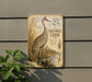 A vintage-style metal sign depicting a sandhill crane standing in a grassy field with tall grass and a body of water in the background.