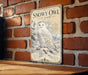 A vintage-style metal sign featuring an illustration of a snowy owl perched on a snowy branch, mounted on a brick wall.
