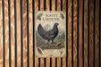 A metal sign with a black and white illustration of a grouse bird, surrounded by text and a decorative border, mounted on a wooden wall.