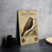 a vintage-style metal sign with a detailed illustration of a star finch, placed on a table next to some black camera lenses.