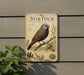 a vintage-style metal sign with a detailed illustration of a Star Finch, a small bird with a distinctive appearance, perched on a branch. The sign is mounted on a wall and is surrounded by greenery, including a plant to the left.