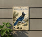 a vintage-style metal sign with a blue jay illustration, mounted on a wall next to a plant.