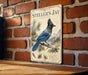 A vintage-style metal sign featuring a blue jay perched on a branch, with text and illustrations related to the bird.