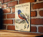 a vintage-style metal sign with a detailed illustration of a colorful bird, likely a species of bird, perched on a branch. The sign is mounted on a brick wall, and there is a wooden table below it.