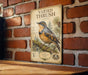 a vintage-style metal plaque with a detailed illustration of a bird perched on a branch, set against a brick wall.