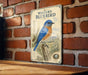 A vintage-style metal sign featuring an illustration of a bluebird perched on a tree branch, with a brick wall in the background.