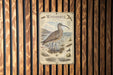 A metal plaque with an illustration of a bird, likely a Whimbrel, standing on a grassy area with a body of water in the background.