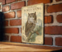 A metal sign featuring an illustration of a Whiskered Screech- Owl perched on a branch, with a brick wall in the background.