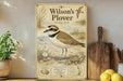 A vintage-style poster of a Wilson's Plover, a small shorebird, is displayed on a shelf next to a wooden cutting board and some potted plants.