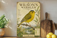 A vintage-style poster of a yellow warbler bird is displayed on a shelf, accompanied by a wooden cutting board and some potted plants.