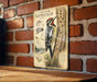 a vintage-style illustration of a yellow-bellied sapsucker, a type of woodpecker, perched on a tree trunk. The illustration includes detailed illustrations of the bird's distinctive features, such as its red crest, black and white stripes, and long, curved beak. The illustration is mounted on a brick wall, and there is a wooden table in the foreground.